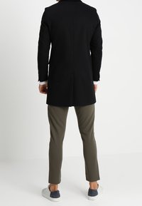 Une personne se tient de dos, portant un manteau noir, un pantalon vert olive et des chaussures sans lacets, devant un fond blanc uni.
