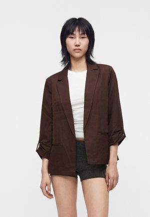 Jeune femme aux cheveux courts noirs portant un blazer marron sur un haut blanc et un short foncé, sur un fond clair uni.