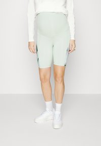 Calças de ciclismo de maternidade em verde menta com uma textura suave, apresentando painéis laterais com um padrão floral. Usadas com ténis e meias brancas.