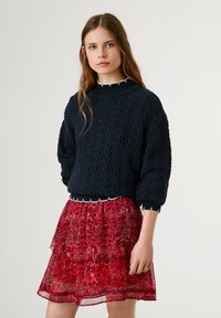 Jūras zilas krāsas adīta džemperis ar ribotu apakšmalu, kombinēts ar caurspīdīgu sarkanu ziedu raksta svārku ar frēzēm un mežģīņu akcentiem.