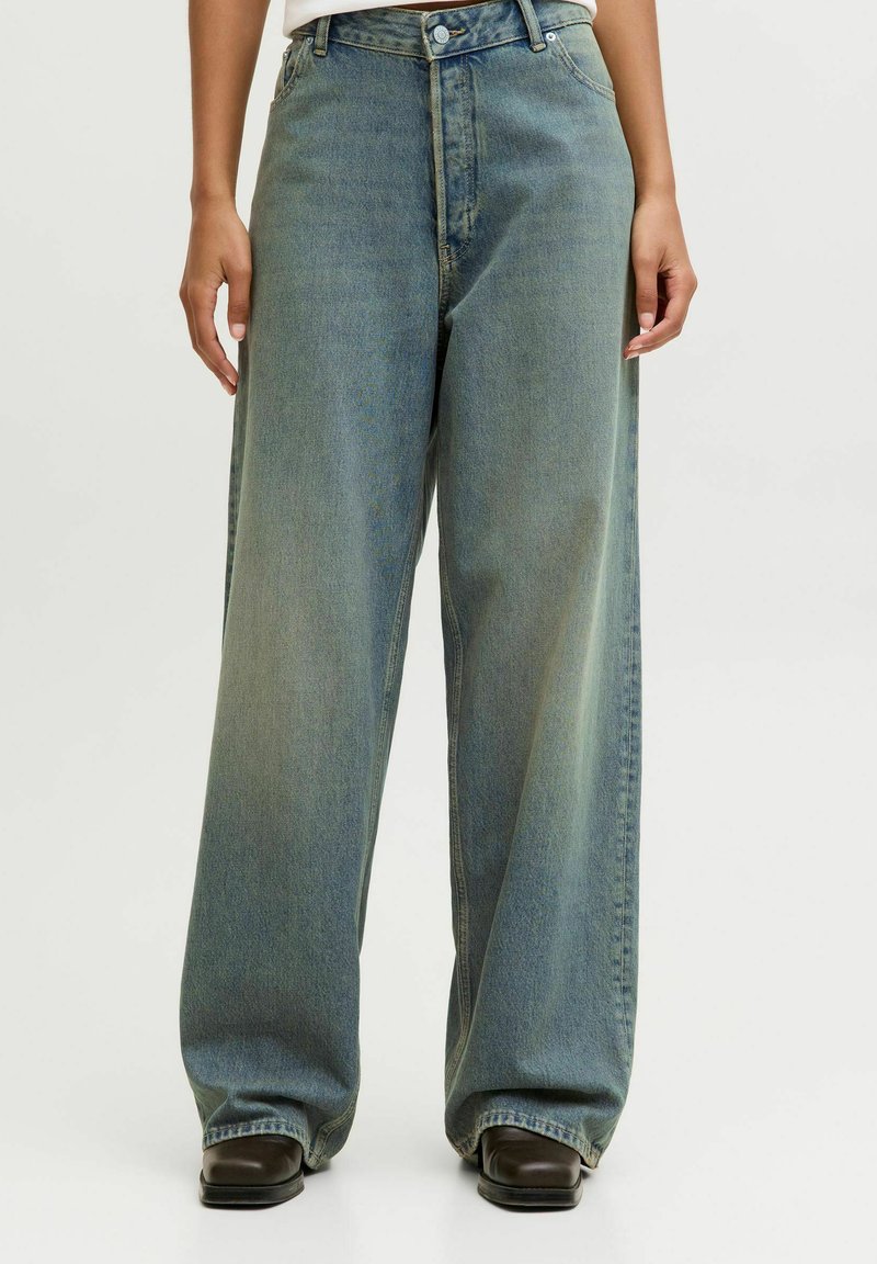 Personne portant un jean bleu ample, taille haute, jambes larges, couvrant des chaussures noires à bout carré, debout avec les mains détendues le long du corps.