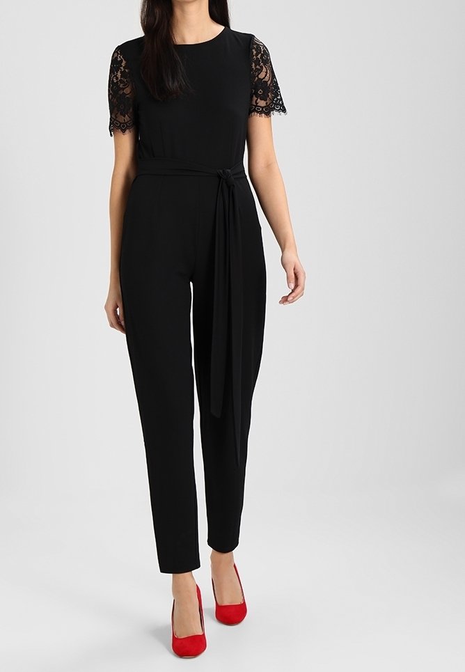 Zwarte jumpsuit met korte mouwen van kant, een ceintuur rond de taille en taps toelopende benen. Gecombineerd met rode hoge hakken voor contrast. Gladde textuur en een aangesloten ontwerp.