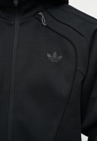 Μαύρο φούτερ με φερμουάρ, με το λογότυπο Adidas trefoil στο αριστερό στήθος και κορδόνια κουκούλας με κλείστρο.