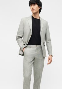 Άνδρας που φοράει ανοιχτό γκρι κοστούμι με κλειστό σακάκι και ταιριαστό παντελόνι, συνδυασμένο με μαύρο μπλουζάκι με στρογγυλή λαιμόκοψη, στέκεται με το ένα χέρι στην τσέπη.