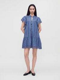 Femme debout, les mains derrière le dos, portant une robe courte en dentelle bleue avec des boutons et des chaussures plates noires sur un fond uni.