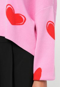 Pink sweater with a relaxed fit, featuring red heart patterns on sleeves. Fabric appears soft; worn over a black garment.