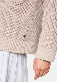 Beige gestrickter Pullover mit gerippten Bündchen und Saum, seitlichen Schlitzen und einem kleinen dekorativen Metallakzent nahe dem Saum.