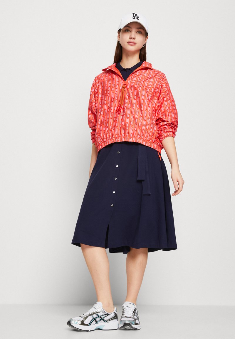 Chaqueta corta con un diseño de rayas rojas y bajo elástico; vestido azul marino con botones y abertura; gorra blanca; zapatillas grises con acentos azules.