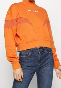 Orange sweatshirt med hög krage, med mörkare orangea böjda detaljer och vit "A+R"-logotyp. Kombinerad med mörkblå jeans.