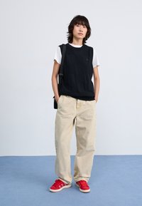 Svart stickad väst över en vit t-shirt, beige lösa byxor och röda sneakers med vit sula. Avslappnad outfit med en texturerad västdesign.