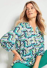 Mehrfarbiges Blusenshirt mit einem geometrischen Muster in Blau, Grün und Schwarz. Lockere Passform mit V-Ausschnitt und umgeschlagenen langen Ärmeln.