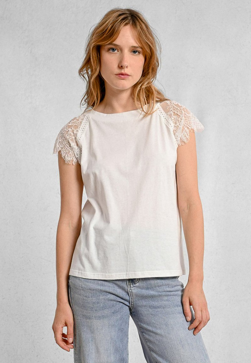 Top in cotone bianco con maniche rifinite in pizzo, dotato di una vestibilità rilassata e scollatura rotonda. Abbinato a jeans azzurri in un look casual.