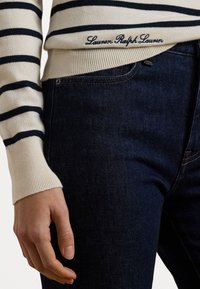 Pruhovaný pletený svetr v krémové a námořnické modré barvě s žebrovaným lemem, kombinovaný s tmavými džínami. Svetr zdobí vyšívané logo značky.