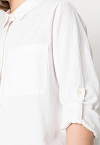 Camisa branca com colarinho abotoado, mangas longas com punhos enrolados, um bolso no peito e um tecido texturizado. Os botões são de cor clara.