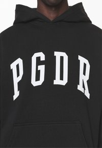 Čierna mikina s kapucňou a predným vreckom, na ktorom sú veľkými bielymi písmenami "PGDR" napísané cez hruď v športovom, veľkými písmenami písanom písme. Mäkká textúra látky.