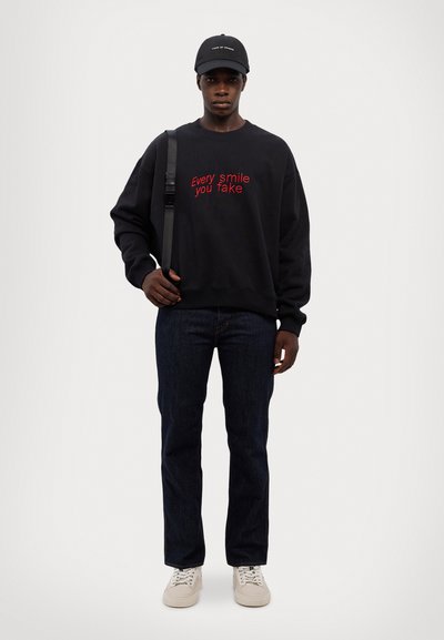 Felpa nera con testo rosso, jeans blu e sneakers beige. La felpa ha una vestibilità ampia; i jeans sono a gamba dritta con un lavaggio scuro.