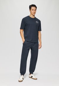 Navyblaues T-Shirt mit kurzen Ärmeln und kleinem Grafikdruck auf der Brust, kombiniert mit passenden navyblauen Jogginghosen und weißen Sneakers mit dunklen Akzenten.