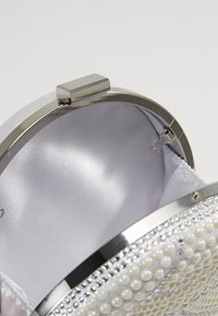 Okrągła srebrna kopertówka z metalową ramą, ozdobiona perełkowymi koralikami i gładkim satynowym wnętrzem. Zapięcie z zawiasem i klamrą.