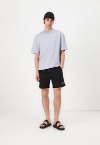 Tricou gri din bumbac cu logo, pantaloni scurți negri cu print pe lateral, sandale negre. Design simplu, croială lejeră, potrivit pentru ținute casual.