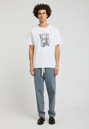 Homme aux cheveux bouclés portant un t-shirt blanc avec un motif ours en peluche, un pantalon rayé bleu avec cordon de serrage et des chaussures noires, debout sur un fond uni.
