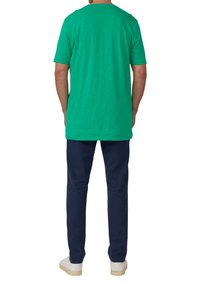 Zielona koszula z krótkim rękawem o luźnym kroju, z okrągłym dekoltem i gładką fakturą, w zestawieniu z granatowymi spodniami i białymi trampkami.