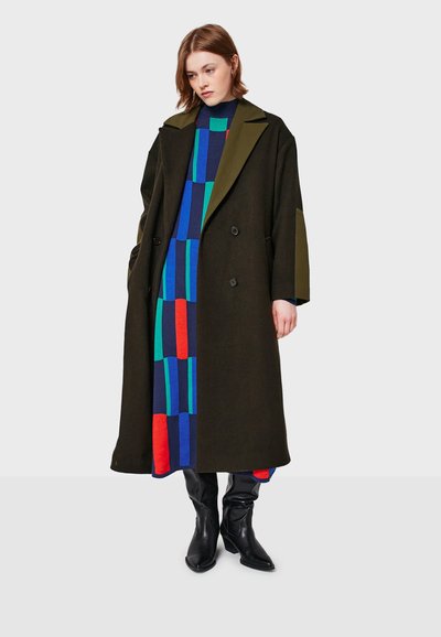 Frau in einem langen dunkelgrünen Mantel über einem bunten Kleid mit blauem, grünem und rotem Muster, kombiniert mit schwarzen kniehohen Stiefeln.