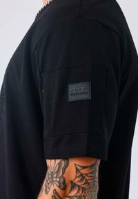 Persona vestida con una camiseta de manga corta negra con un logo de PX-P Connexion en la manga, mostrando un antebrazo tatuado con diseños de mariposa y telaraña.