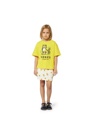 T-shirt di cotone gialla con stampa di tigre e testo "KENZO PARIS", abbinata a una gonna bianca con motivi di girasoli gialli. Scarpe nere.