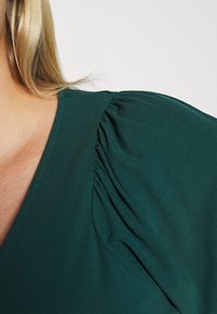Vihreä ribattu kankainen paita, jossa on V-pääntie, kerrokselliset puffihihat ja sileä pinta, korostaen olkapään lähikuvaa.