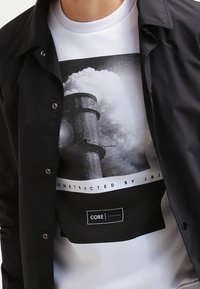 Schwarze Snap-Button-Jacke über einem weißen Sweatshirt mit einem großen schwarz-weißen grafischen Druck von industriellen Schornsteinen und einem Textlabel.