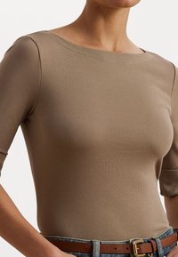 Top castanho ajustado feito de um tecido suave, com decote barco e mangas curtas, combinado com jeans azuis de cintura alta e um cinto castanho.