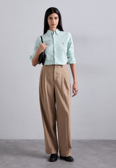 Camicia a righe verde chiaro con bottoni, pantaloni beige a vita alta con pieghe e scarpe nere; tiene in mano una borsa nera. Tessuti con texture visibili.