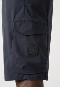 Shorts bleu marine en tissu lisse et léger, dotés d'une poche cargo et d'une étiquette rectangulaire noire avec du texte vert.