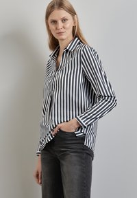 Camicia a righe blu navy e bianca, con maniche lunghe, colletto e un leggero strascico con spacchetti laterali, abbinata a jeans scuri.