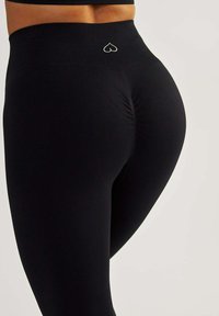 Leggings noirs taille haute en tissu extensible, présentant un design froncé à l'arrière et un petit logo en forme de cœur sur la ceinture.