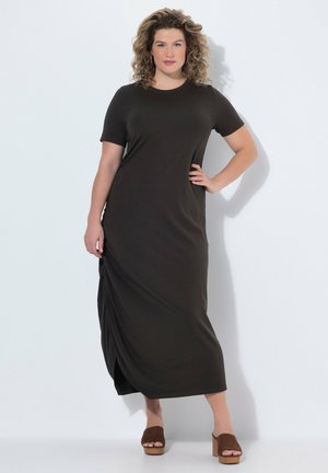 Žena s kudrnatými vlasy stojí s rukou na bocích, oblečená v černých midi šatech s krátkým rukávem a hnědých platformových sandálech na bílém pozadí.