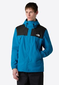 Modrá a černá větrovka s kapucí, s elastickými manžetami, zapínáním na zip v přední části a logem na hrudi. Hladká, lehká tkanina.