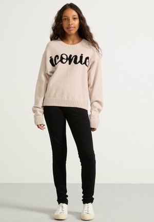 Ljusrosa tröja med "iconic" i svart skrift, ribbat nederkant och ärmslut; matchad med svarta skinny jeans och vita sneakers.