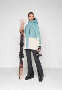 Ski jacket in teal and cream, black pants, holding skis with bindings. Features a hood, zippered pocket, and ski goggles in hand.