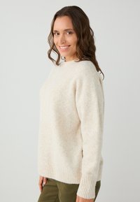 Pull en tricot beige avec une texture douce, col rond et poignets côtelés. Porté sur un pantalon vert olive, présenté sur un fond neutre.