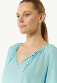 Light blue blouse made of soft, flowing fabric. Features a V-neckline with gentle pleats and long, relaxed sleeves.