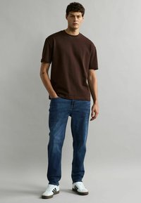 Camiseta de algodón marrón de mangas cortas, combinada con jeans de mezclilla azul. El modelo lleva zapatillas blancas con rayas negras y está de pie de manera casual.