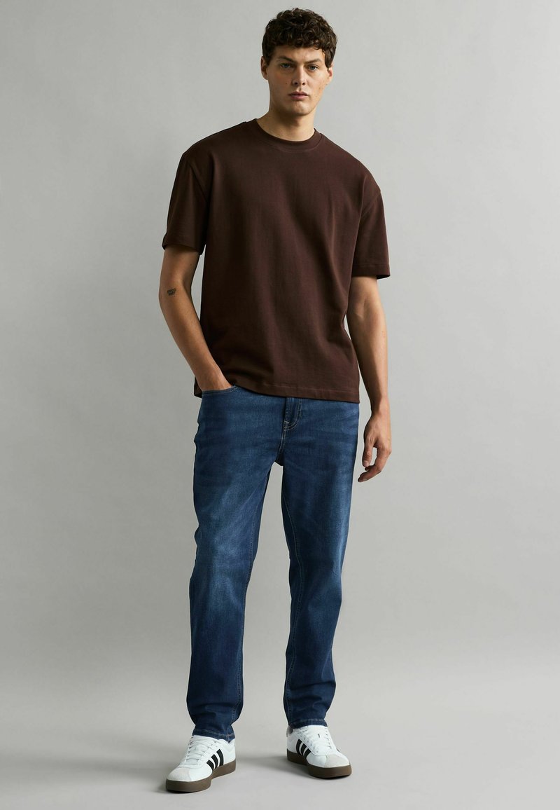Camiseta de algodón marrón de mangas cortas, combinada con jeans de mezclilla azul. El modelo lleva zapatillas blancas con rayas negras y está de pie de manera casual.
