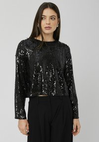 Haut noir à manches longues raccourci en tissu pailleté, avec un design à rayures texturées et un col rond. Porté avec un pantalon taille haute.