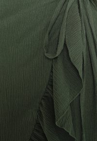 Grüner, strukturierter Stoff mit weichen, zerknitterten Falten und subtilen Drapierungselementen, die Tiefe und Bewegung im Design erzeugen.