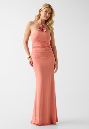 Donna con lunghi capelli biondi che indossa un abito senza maniche color corallo rosa con una decorazione floreale sulla scollatura, in piedi davanti a uno sfondo semplice.