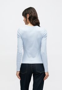 Jasnoniebieska koszula z długim rękawem z prążkowanej tkaniny, z okrągłym dekoltem i dopasowanym krojem. Noszona z ciemnymi dżinsami. Widok od tyłu.