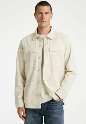 Jonge man met kort haar die een beige overhemd met knopen draagt over een licht t-shirt en blauwe spijkerbroek, staand tegen een effen achtergrond.