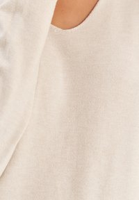 Beige stickad tröja med v-ringad design och mjuk textur. Tyget verkar slätt med subtila nyansvariationer.