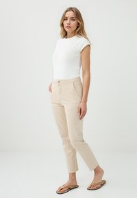 Jonge vrouw staat zelfverzekerd in een witte getailleerde top en beige broek, met bruine slippers aan, tegen een effen lichte achtergrond.